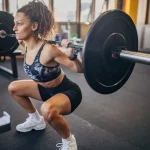 Treino Full Body Feminino 3x na Semana (Ficha Completa em PDF) Mulher fazendo agachamento livre com barra, ativando glúteos, quadríceps e isquiotibiais.