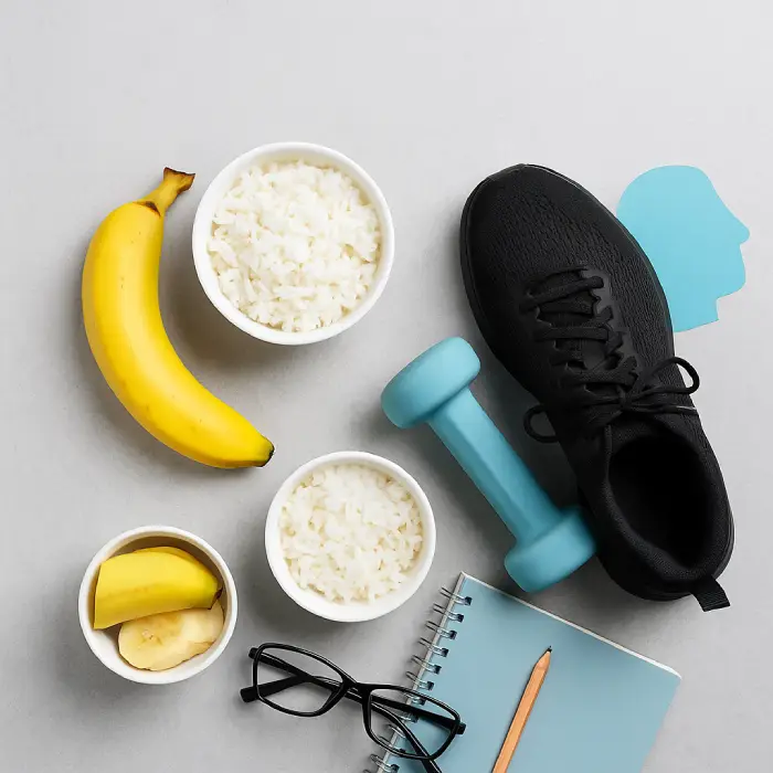 Banana, tigelas com arroz branco, halter azul claro, tênis preto, caderno, lápis e óculos organizados sobre superfície cinza clara, simbolizando a relação entre nutrição e treino físico