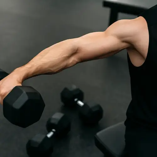 Close no braço de homem realizando elevação lateral com halteres pretos, destacando ombro e antebraço em contração com fundo desfocado de academia