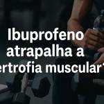 Corpo de atleta sentado em banco de academia segurando garrafa d’água, com halteres no chão ao redor. Texto sobreposto: 'Ibuprofeno atrapalha a hipertrofia muscular?